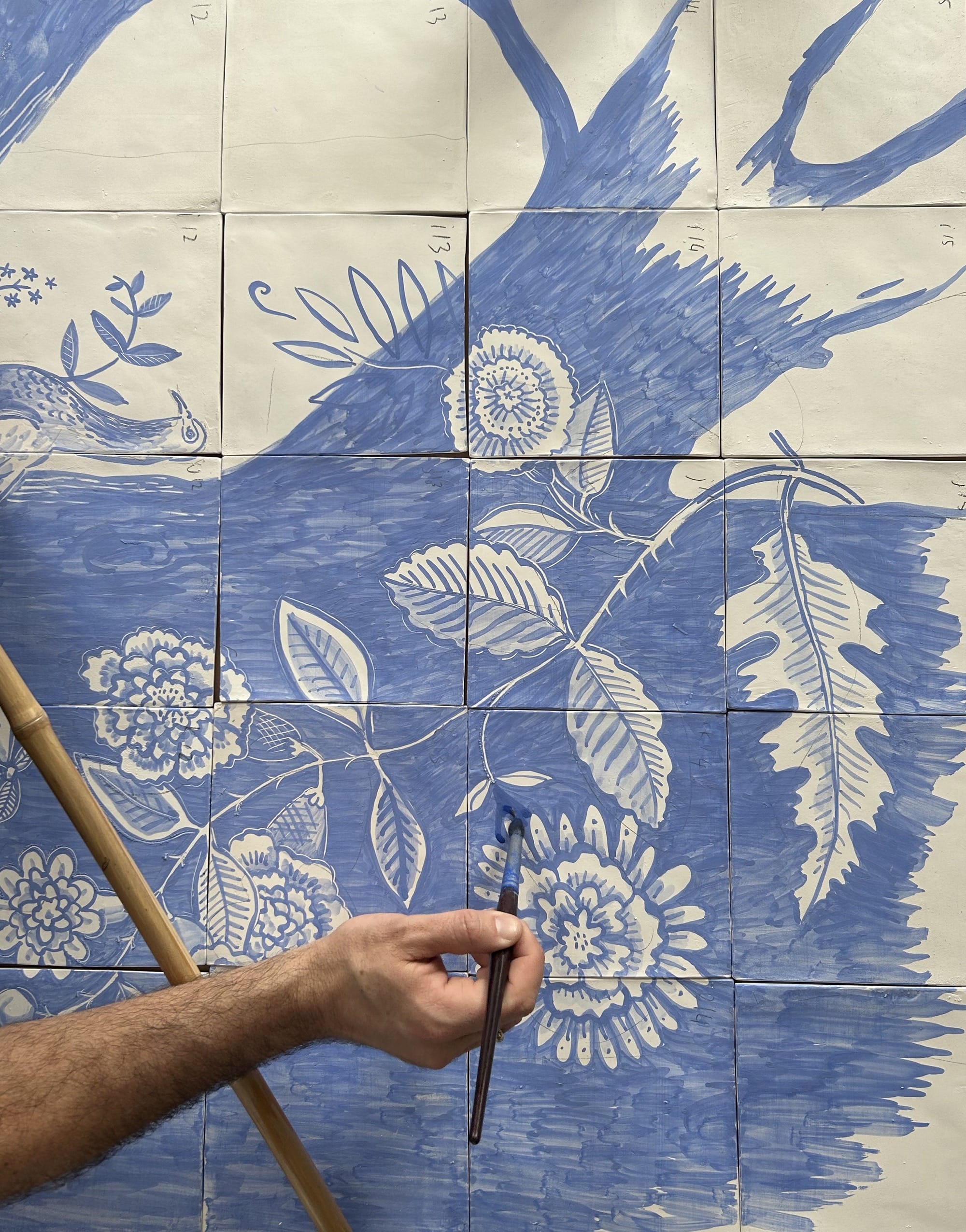 a hand paints flowers in blue glaze onto white ceramic tiles