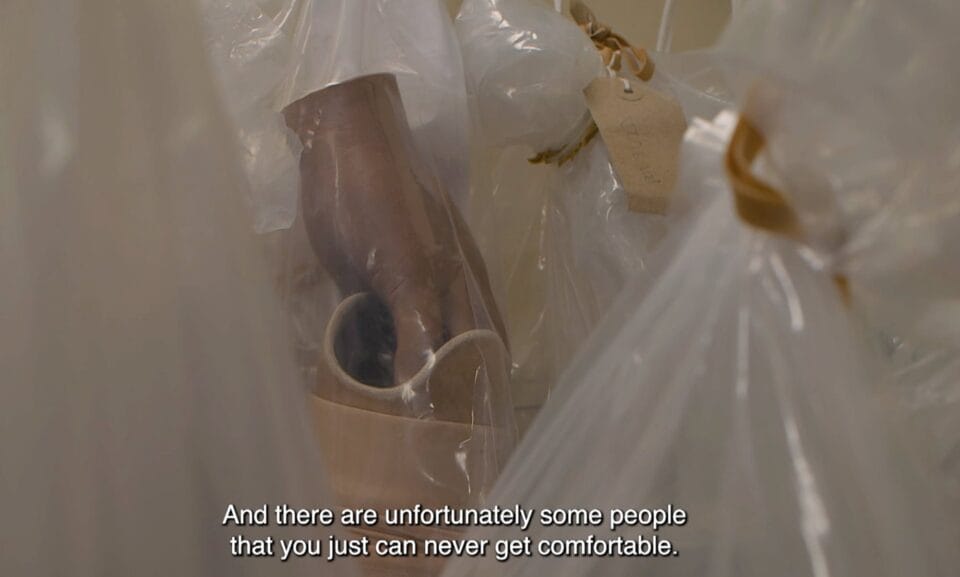 a video still of plastic bags holding prosthetics with a prosthetic hand and leg in one of the bags. a caption reads "and there are unfortunately some people that you just can never get comfortable."