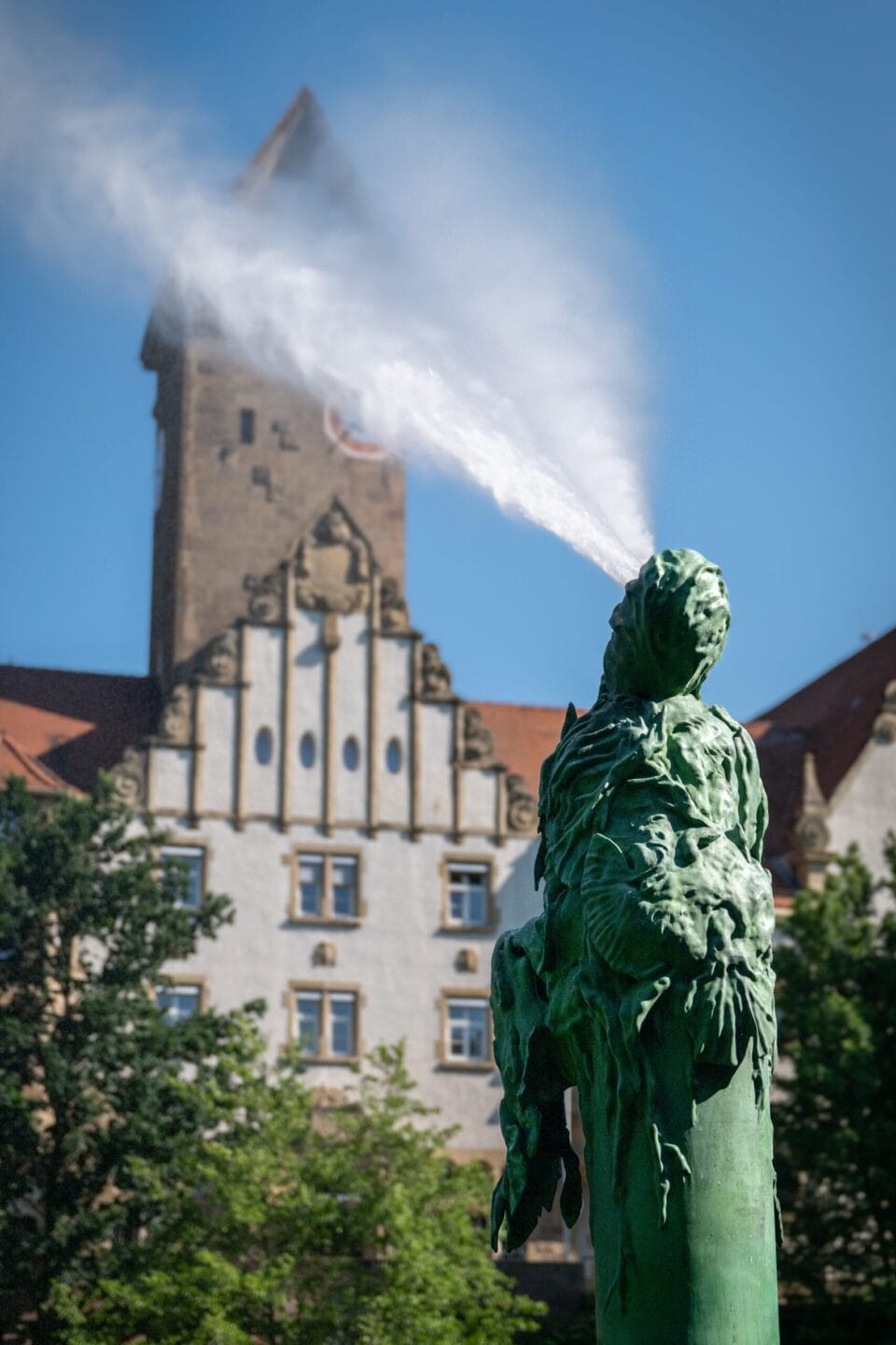 a tall fountain that resembles a sitting elf-like figure is situated next to a river as it sprays water out of its head