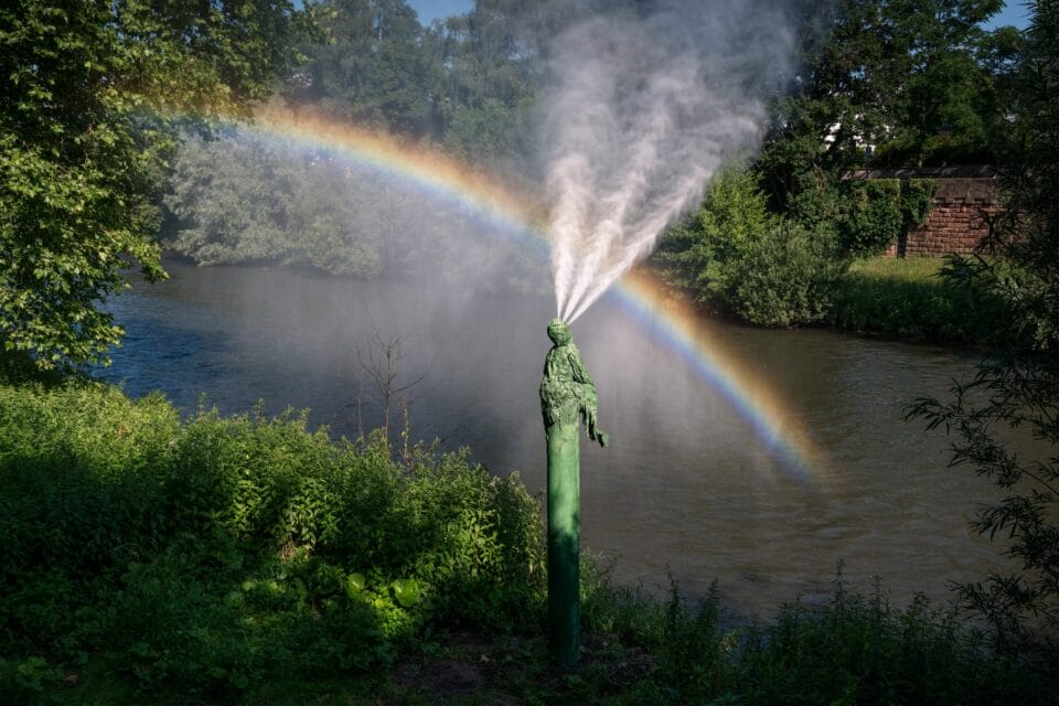 along a river, a fountain sprays water as a rainbow appears next to it