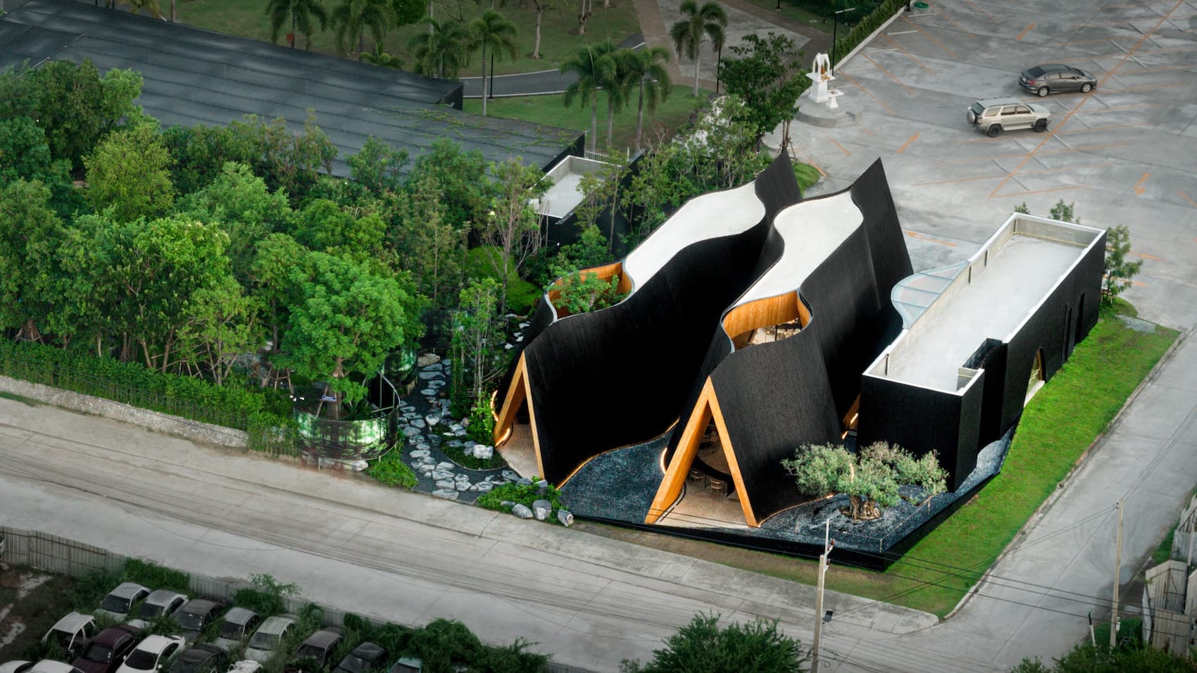 an aerial view of a contemporary coffee shop in Thailand featuring two A-frame gables, undulating black sides, and voids in the ceiling in two places to support trees growing indoors