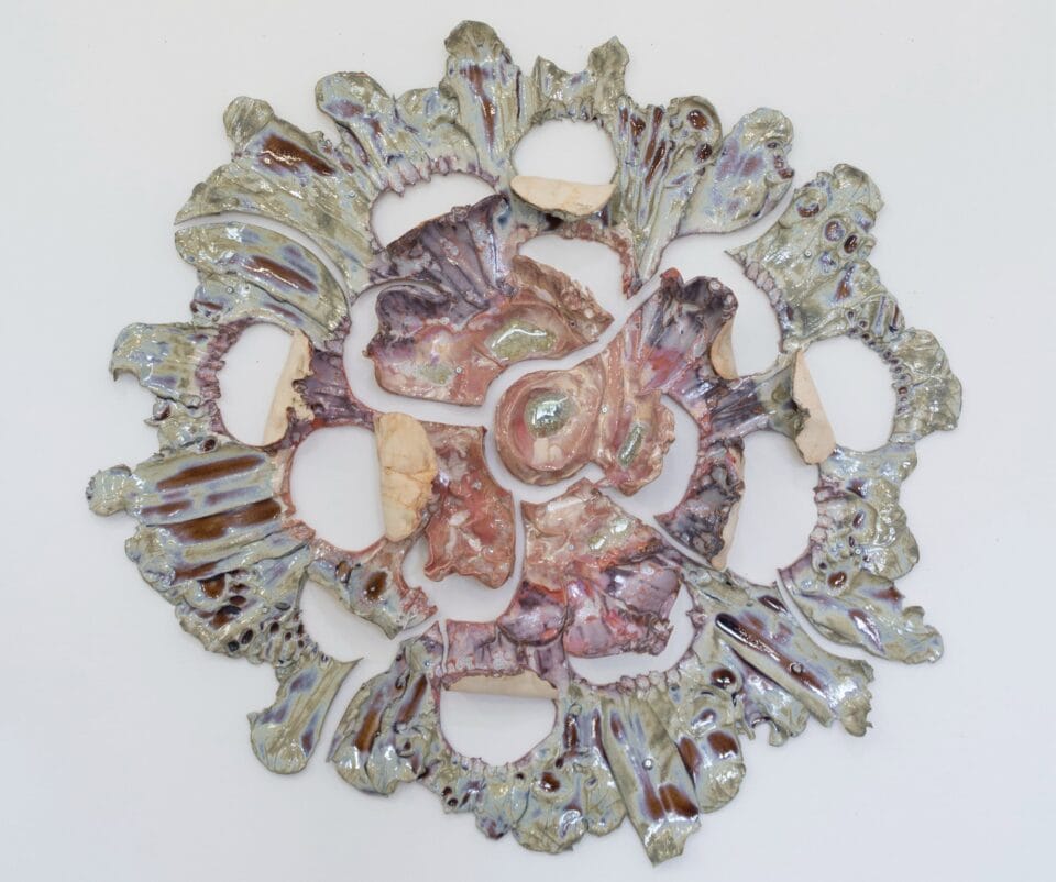 a round pink and green -gray wall sculpture that shows pieces of ceramic with lines evocative of stretching radiating from a central point. 