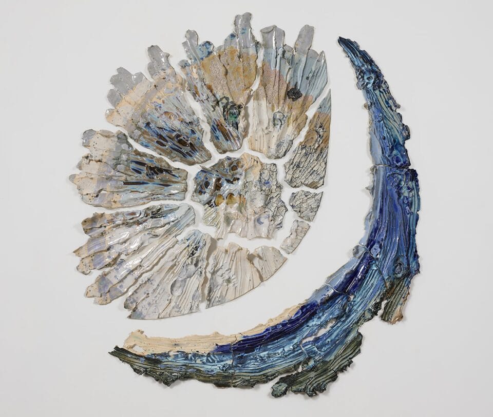 a round beige and white wall sculpture that shows pieces of ceramic with lines evocative of stretching radiating from a central point with the right and bottom sides cut cleanly. a curved blue piece cradles the right and bottom sides