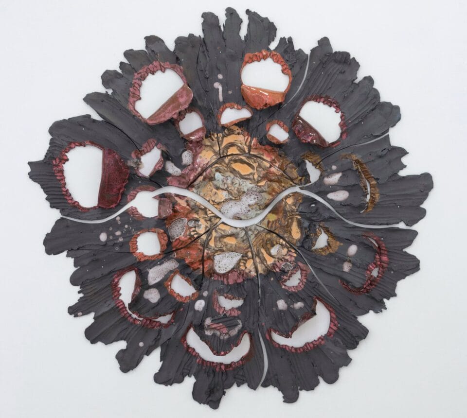 a round pink, gray, and orange wall sculpture that shows pieces of ceramic with lines evocative of stretching radiating from a central point. a squiggly crack runs horizontally down the center