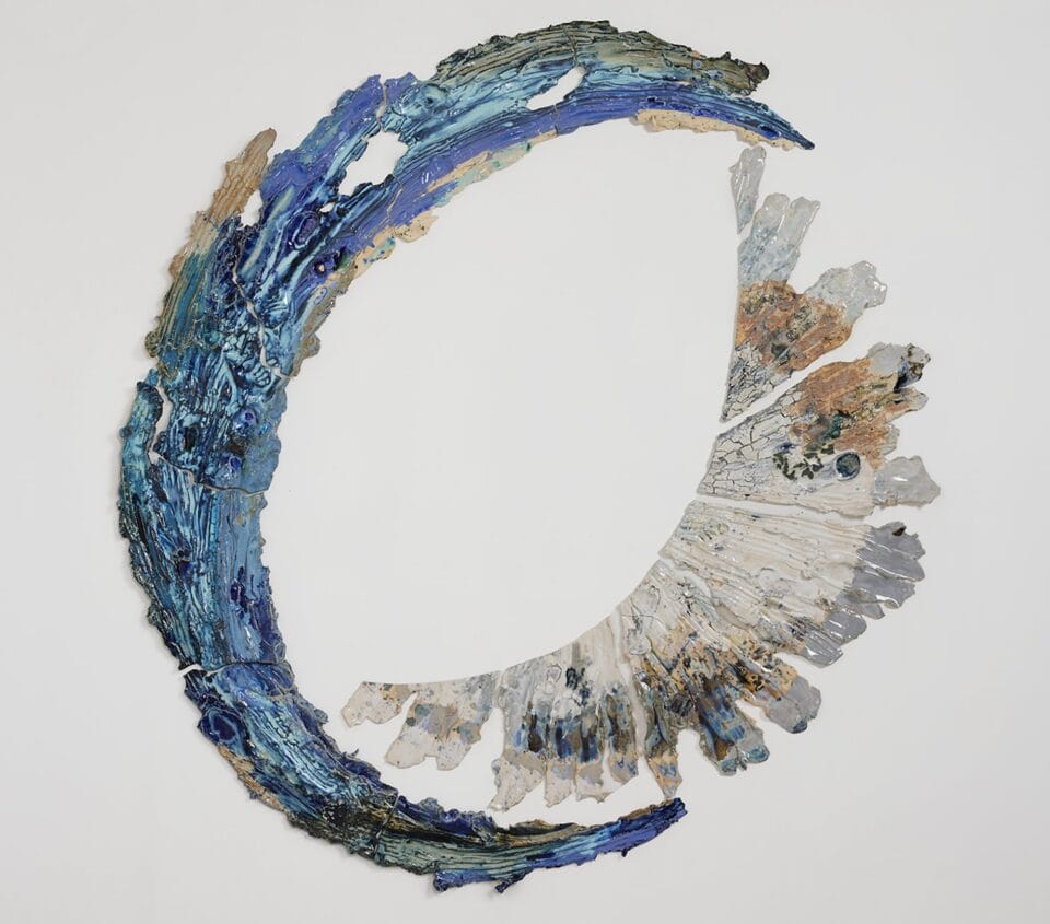 a round beige and white wall sculpture that shows pieces of ceramic with lines evocative of stretching radiating from a central point with the left and bottom sides cut cleanly. a curved blue piece cradles the left and bottom sides