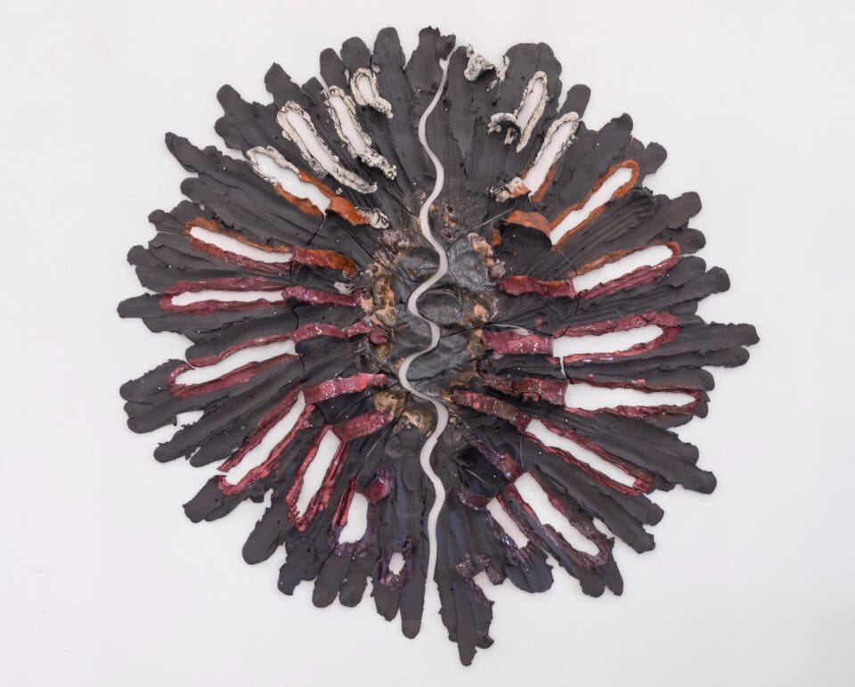 a round pink, gray, and orange wall sculpture that shows pieces of ceramic with lines evocative of stretching radiating from a central point. a squiggly crack runs vertically down the center