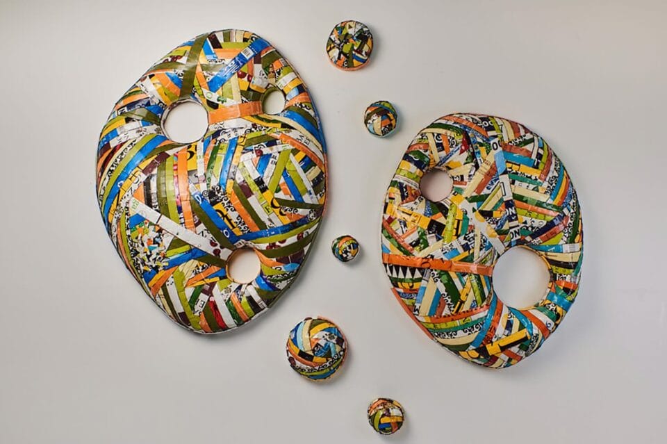 an abstract cardboard wall sculpture with two main round pieces and five smaller satellite pieces, predominantly blue, green, orange, white, and brown, made from found boxes cut into strips