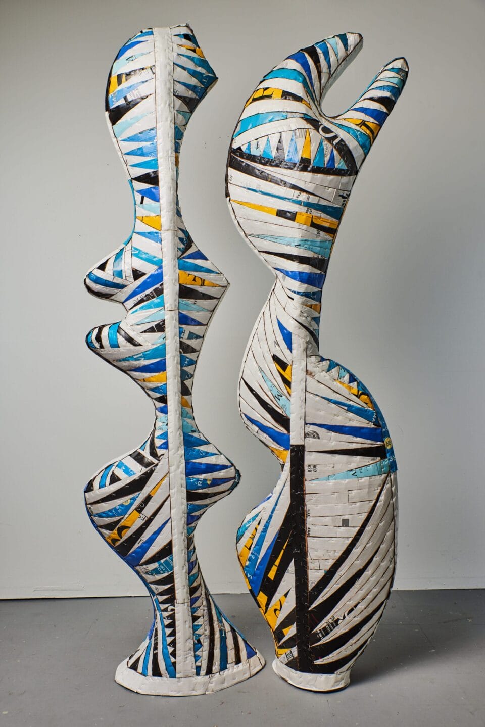 two freestanding sculptures side-by-side made of cardboard that has been cut into strips and layered, primarily in white, blue, black, and yellow