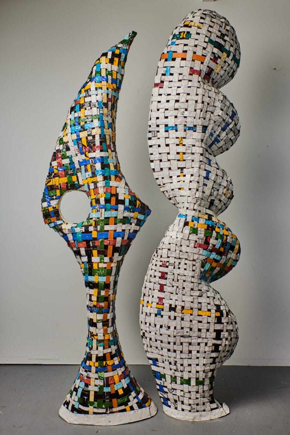 two freestanding sculptures side-by-side made of cardboard, the one of the right predominantly a white woven structure with spots of color, and the one of the left mostly colorful with spots of white